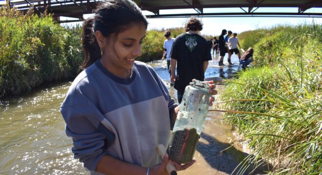 A young girl smiles down at a water bottle filled with river sediment as students wade through a river behind her.