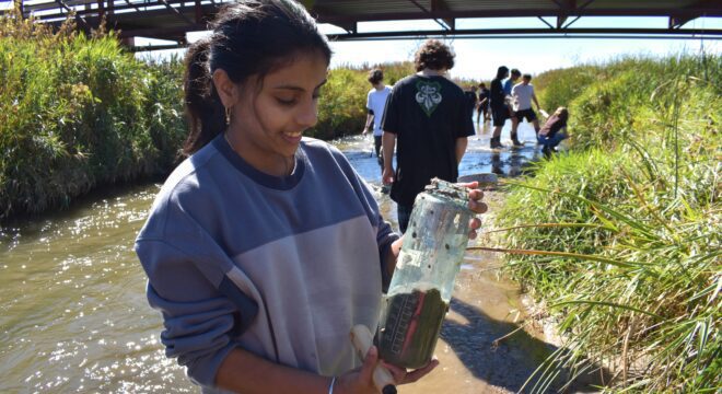 A young girl smiles down at a water bottle filled with river sediment as students wade through a river behind her.
